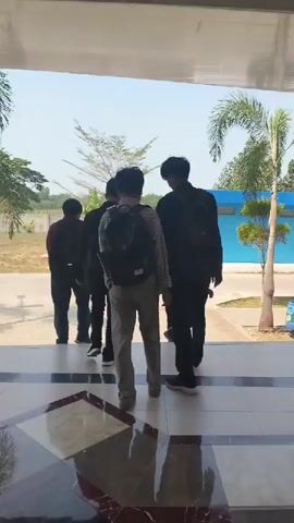 The video begins with a view from inside a building looking outwards through an entranceway. Four individuals, all appearing to be male, are walking away from the camera and down a set of steps. They are all wearing backpacks and dark-colored clothing, except for one person in the center who is wearing a light-colored patterned shirt and tan pants. The environment outside appears to be a sunny day with a paved walkway, grass, trees, a blue building in the background, and a palm tree to the right. The camera is positioned low, giving a perspective from floor level looking up at the individuals as they descend the steps and exit the building.