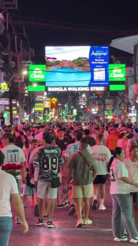The video captures a bustling night scene on a crowded street, illuminated by numerous artificial lights and large digital billboards. The street is packed with a diverse crowd of people walking in various directions, suggesting a popular entertainment or shopping district.

Dominating the upper portion of the frame are large, brightly lit digital billboards. A prominent central screen displays advertisements, cycling through scenes of people enjoying a water park and aerial views of a resort. Text on this screen reads "ANDAMANDA The Great Water Kingdom" along with a QR code and the website "www.andamandaphuket.com". Below this, a large sign clearly spells out "BANGLA WALKING STREET". Flanking the central screen are two smaller, rectangular green billboards, each displaying the white "Chang" beer logo.

The street itself is wide and appears to be a pedestrian-only zone, as no vehicles are visible. Overhead, a dense network of electrical wires and cables can be seen. Buildings lining both sides of the street are adorned with various signs and neon lights, though specific details of most establishments are obscured by the crowd and distance.

The people in the video are casually dressed in t-shirts, shorts, and some in long pants, indicative of a warm climate. Many are walking and mingling, while some appear to be looking at their phones. Several individuals are seen carrying large bouquets of red roses, particularly prominent are two people in the foreground on the left and right. One man, walking away from the camera, wears a jersey with "DESANYA 89" printed on the back. Towards the end of the clip, a man walks across the foreground from left to right, pushing a small, rectangular cart that appears to hold some items or a game.

The atmosphere is vibrant and energetic, consistent with a busy night street. Upbeat, somewhat distorted music can be heard playing in the background, adding to the lively ambiance.