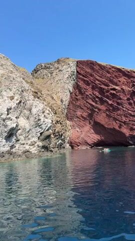 A bright, clear day unfolds with a striking geological feature of a cliff face meeting the sea. The cliff is divided by color: the left side is a pale, almost white rock, while the right side is a vibrant, deep red. This division creates a dramatic visual contrast against the azure sky and the deep blue of the water. The water itself is calm, with ripples reflecting the sky and the cliff face. A small, light-colored boat, carrying at least three people, is positioned near the base of the red cliff, suggesting they are exploring or enjoying the scenery from the water. The overall impression is one of natural beauty and a serene maritime environment.