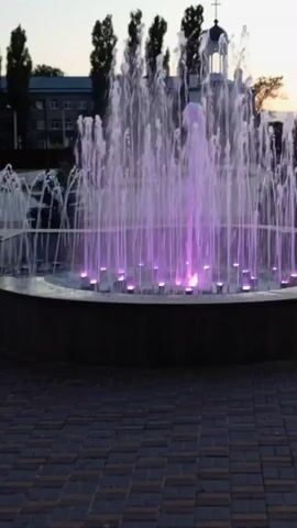 The video shows a large, illuminated fountain in what appears to be a public space. The fountain has multiple streams of water shooting upwards and outwards, creating a dynamic display. At the base of the fountain, there are colored lights that illuminate the water, cycling through various hues of blue, green, pink, and yellow. In the background, there are trees and a building with a cross on its dome, suggesting it might be a church or a similar institution. The ground in front of the fountain is paved with bricks arranged in a pattern. The sky above appears to be a pale twilight or dawn sky. The video is a continuous shot of the fountain without any discernible action or interaction from people.