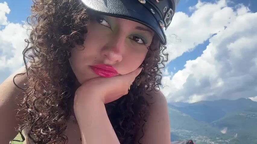 This close-up media file features a young woman with curly brown hair and dramatic green eyeshadow. She is wearing a black hat, possibly a visor, and has bright pink lipstick. Her right hand is resting on her chin, supporting her head as she looks slightly off-camera. Sunlight casts dappled shadows across her face.

The background suggests an outdoor setting on a sunny day with puffy white clouds against a blue sky. Mountains are visible in the distance, and what appears to be a body of water, possibly a lake, is also present. In the far left background, a person in swim trunks stands near the water, and some beach chairs or towels are visible on the grassy area. The overall impression is one of a relaxed, outdoor leisure activity, likely in a mountainous region with a lake.

There is no visible text in the media file. The time of day appears to be daytime, given the bright sunlight and clear sky. The weather is fair and sunny, with a gentle breeze suggested by the movement of the woman's hair and the clouds. The location context provided is Colico, Italy, which aligns with the mountainous landscape and the presence of a lake. The woman's pose and expression could be interpreted as contemplative, relaxed, or perhaps a bit bored, but without further context, it's difficult to ascertain a specific emotion.
