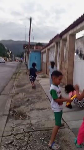 The video shows a group of children playing outside on a sidewalk in a residential area. There are at least four children visible, running and interacting playfully. A child wearing a white and green outfit is initially in the foreground, then turns and runs after the others. The other children are wearing various casual clothes. They are running down a sidewalk next to a row of houses. The houses are made of beige or tan brick and some have white metal fences.  A street with vehicles is visible in the background, with mountains in the distance under a cloudy sky. The overall environment suggests an urban or suburban setting. Sounds of children and barking dogs are heard in the background.
