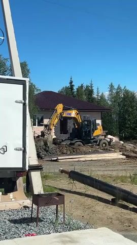 A yellow excavator is digging a hole next to a house. The excavator is in the process of removing dirt from the ground. The house is a two-story building with a brown roof. There are trees in the background. A white truck is parked to the left of the frame. There is also a black pipe lying on the ground in the foreground. The sky is blue and clear.