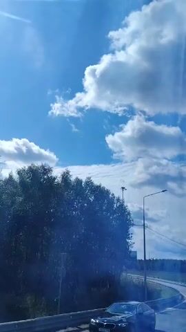 A video taken from a moving vehicle shows a clear blue sky with fluffy white clouds. The road curves to the right, bordered by a metal guardrail and trees on the left. Streetlights are visible along the roadside. The sun is shining brightly, casting lens flares.