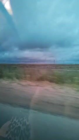 The video shows a view from inside a moving vehicle. The perspective is through the car window, offering a slightly blurred but discernible scene of a flat landscape. The sky is predominantly filled with dark, ominous clouds, suggesting an impending storm or overcast weather. A line of wind turbines is visible on the horizon, spaced out across the flat terrain. The ground in the foreground appears to be a mix of dirt and vegetation, also blurred due to the motion of the vehicle. In one segment, the interior of the car is visible, showing a person in the passenger seat looking out the window. The overall impression is one of travel through a wide-open, somewhat desolate landscape under a dramatic sky.