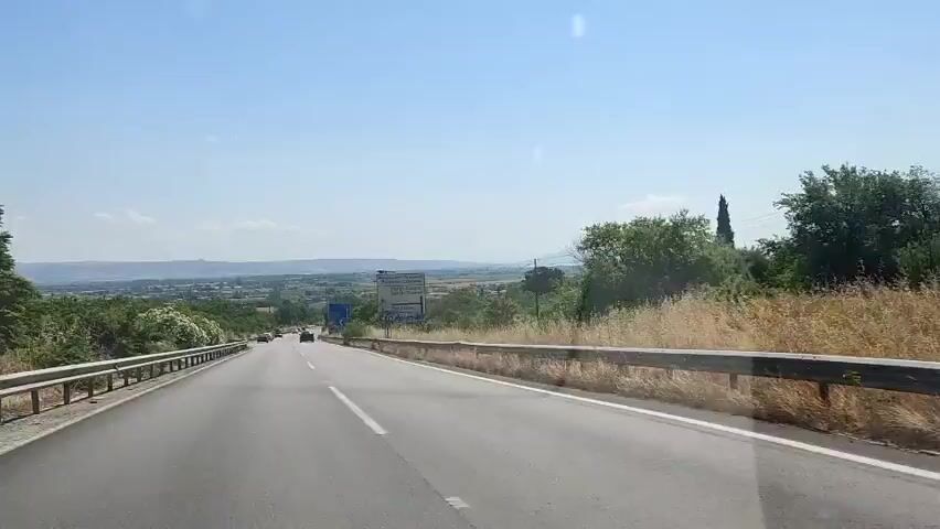 The video captures a first-person perspective from a vehicle driving on a paved road under a clear, bright sky. The road has two lanes marked with white lines and is flanked by metal guardrails on both sides. Sparse green and dry vegetation lines the road. In the distance, a rural landscape with scattered buildings and distant mountains is visible under a hazy sky. Several cars are seen driving in the distance, and utility poles with power lines run alongside the road. A large white sign with text in multiple languages, including what appears to be Bulgarian and Greek, stands on the right side of the road, reading "Bulgaria - Customs." Another blue sign is also visible further down the road on the right.
