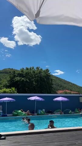 The video pans across a swimming pool on a sunny day. The pool is surrounded by a wooden deck and a blue wall adorned with purple umbrellas and green beanbag chairs. Several people are in the pool, some swimming and others relaxing. In the background, a lush green mountain rises against a clear blue sky with white clouds. The overall atmosphere appears to be one of leisure and relaxation.