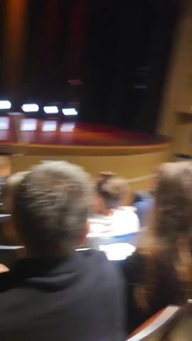 The video shows a crowd of people seated in an auditorium, facing a stage. The lighting in the auditorium is dim, with spotlights illuminating the stage. The audience members are mostly blurred, but some can be seen looking towards the stage. The stage is set with a dark backdrop and a few lights. The overall atmosphere appears to be one of anticipation, as if an event is about to begin.