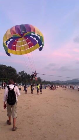 The video shows a scene on a sandy beach with people parasailing. A large, colorful parachute is visible, held up by several ropes connected to a group of people being prepped for the activity. Other people on the beach observe the parasailing preparation. A man wearing a white shirt, light-colored shorts, and a backpack walks in the foreground. The sky displays hues of pink and light purple, suggesting it might be dusk. There are trees and hills in the background. Beach umbrellas and lounge chairs are also present in the scenery.