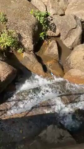 The video shows a small stream of water flowing over and around a collection of rocks. The rocks are various shades of gray and brown, and some have small green plants growing on them. The water is clear and bubbly as it rushes over the rocks, creating small whitecaps. The camera angle is a high-angle shot, looking down on the stream. Light is dappled across the scene. There is some visible pollution: a blue plastic item is seen trapped in the rocks.