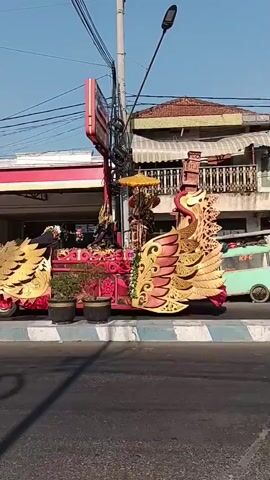 A parade float resembling a large golden bird with red accents is on the side of a street. The float is decorated with red and gold ornamentation and has potted plants. A person in dark clothing with a backpack is walking behind the float on the left side. In the background, a convenience store with red and white signage is visible, along with a street lamp and utility wires. A green food cart with "KFC" visible is also parked further down the street. The sky is clear and blue.