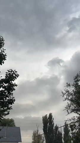This is a vertical video of a cloudy sky from an outdoor perspective. The clouds are dark and heavy, suggesting an overcast or stormy day. Silhouettes of trees and a rooftop are visible in the foreground, framing the sky. A power line stretches across the middle ground, adding an urban element to the natural scene. The overall mood is somber and perhaps a bit foreboding due to the heavy clouds. The video appears to be taken during the daytime, though the lack of direct sunlight and the dense cloud cover make it difficult to pinpoint the exact time. There are no discernible people or specific objects that suggest a particular activity or event. The location context provided is Ават, Kazakhstan, which adds geographical information to the otherwise general outdoor scene.
