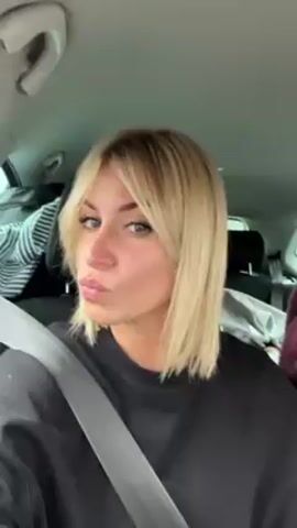 A woman with blonde, shoulder-length hair and bangs is seated in the driver's seat of a car, wearing a black sweatshirt and a seatbelt. She is looking at the camera with a slight pout. The car's interior is visible, with clothing items in the back seat.