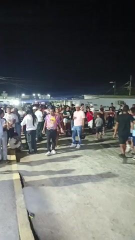 The video shows a crowd of people gathered outside at night. They are standing and walking around on a paved area with buildings and streetlights in the background. Some appear to be talking to each other. The video is shaky and the image is somewhat blurry.