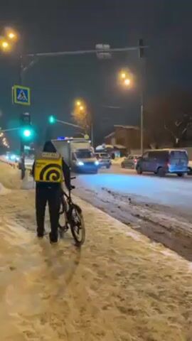 The video shows a delivery person in a yellow and black uniform and backpack walking along a snow-covered sidewalk next to a city street at night. The person is pushing a bicycle. There are several cars on the street, with a green traffic light visible above. Streetlights illuminate the scene. A pedestrian crossing sign is also visible.  The road surface is wet and covered with snow.