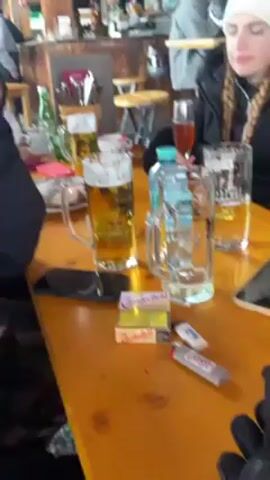 The video shows a group of people sitting at a table outside at what appears to be a ski resort bar.  There are mugs of beer, a wine glass, and a water bottle on the table. There are cigarettes and lighters as well. A man in a fur hat takes a sip from a wine glass. Outside, people in ski gear are walking around the snowy patio area, which has green umbrellas.