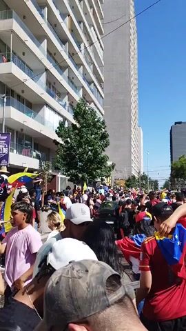 The video shows a large crowd of people, many waving Venezuelan flags, moving down a city street. The scene is sunlit with a clear blue sky. Tall buildings line the street, including a modern high-rise apartment building and an establishment with an unknown black sign. People wear a variety of clothing, with many sporting caps or the colors of the Venezuelan flag. Some people are holding drinks. The crowd appears to be participating in a festive or celebratory gathering.
