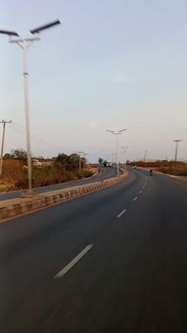 The video shows a straight, winding road in a rural area. The road is paved and has painted white lines marking the lanes. There are streetlights lining the road. On the left side of the road, there is a concrete barrier. Beyond the barrier, there is some vegetation. On the right side of the road, there are no barriers but some vegetation. A motorcycle is visible on the road ahead, and a truck is further down the road. The sky is clear and pale blue. The sound of the wind is noticeable throughout the video.