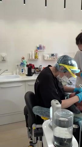 The video shows a dental procedure in progress. Two dental professionals are attending to a patient. The primary dental professional, wearing a black scrub top, a surgical cap, a mask, and a face shield, is seated and actively working on the patient's mouth with dental instruments. The second dental professional, wearing a white t-shirt and a mask, is standing and assisting, holding a light and possibly another instrument. The patient is lying back in a dental chair, wearing protective eyewear. The environment appears to be a clean, well-lit dental operatory, with medical equipment and supplies visible in the background, including a sink, cabinets, and various dental tools. The focus is on the meticulous work being performed by the dental team.