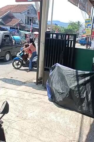 A woman in a pink shirt and blue jeans is sitting on a blue and white motorcycle. She is facing right and appears to be talking to someone behind her. A person in a brown shirt and black pants is also on the motorcycle, sitting behind the woman. They are in a street with other vehicles and buildings. A black pickup truck is driving past on the left. There are food carts and a white SUV in the background. The ground is paved, and there is a black gate on the right. The scene is outdoors during the day.