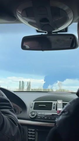 From within a moving car, the dashboard is visible with the steering wheel held by someone wearing a dark jacket.  The rearview mirror reflects the back of the driver's head.  Through the windshield, a large plume of dark smoke rises in the distance against a partly cloudy sky. Trees and utility poles dot the landscape. The car appears to be driving on a highway.