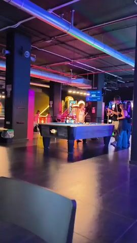 A group of four people are in a dimly lit entertainment venue with neon lights and a reflective floor. In the foreground, a pool table is visible with several balls and cues on it. A woman in a white tank top is standing at the pool table, holding a cue stick, and appears to be playing pool. To her right, two other women, one in a black strapless top and the other in a black jacket, are standing and holding cue sticks. A man in a black shirt and jeans stands behind them. The background shows arcade games with colorful lights. The time on a digital screen in the background reads 00:25.