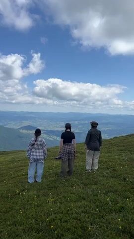 Three people are lying on their backs in a grassy field with their legs in the air. The person on the left is wearing a plaid shirt and blue jeans. The person in the middle is wearing a black t-shirt and dark pants. The person on the right is wearing a hat and a green shirt. The background is a scenic view of rolling hills and a blue sky with white clouds.
