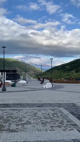 A low-angle vertical video shows a paved walkway leading towards a mountain range. The walkway is made of grey and black interlocking pavers, with a patterned border in the foreground. Several modern streetlights with dark poles and minimalist white fixtures are spaced along the walkway. White benches with rounded edges are placed on either side of the path. In the mid-ground, a few people, appearing to be tourists with luggage, walk away from the camera down the path. A road curves into the distance on the left side of the frame, bordered by green, grassy hills. The background is dominated by a majestic mountain range with snow-capped peaks, partially obscured by dramatic, grey and white clouds. The sky above is a mix of blue and cloudy.