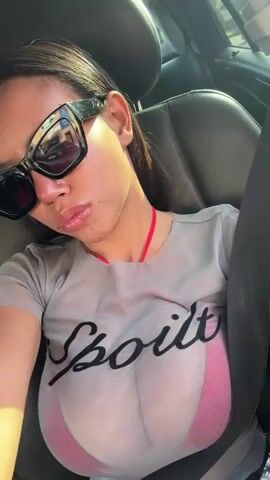 A woman is featured in a close-up selfie video taken inside a car during the daytime. She is wearing large, dark sunglasses and a light grey, sheer, short-sleeved top over a pink bra. The word "Spoiled" is printed in black script across the front of her top. She has a slight smile and appears relaxed, briefly touching her hair or sunglasses. The car's interior, including a dark car seat and the seatbelt across her chest, is visible. The background through the car window is blurred, indicating movement or a bright outdoor setting, consistent with sunny weather in Botany, Australia.