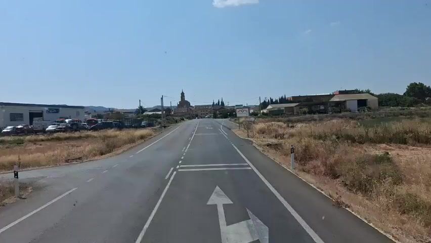The video shows a POV shot from a vehicle driving on a paved road towards a small town under a clear blue sky. The road has white lane markings and a crossing with white parallel lines. On the left side of the road, there is a parking lot with several cars and a building with the text "LAUREL" visible. On the right side, there are dry grasses and some signs. The town in the distance has buildings, including a church with a tall tower. The overall environment appears rural and arid.