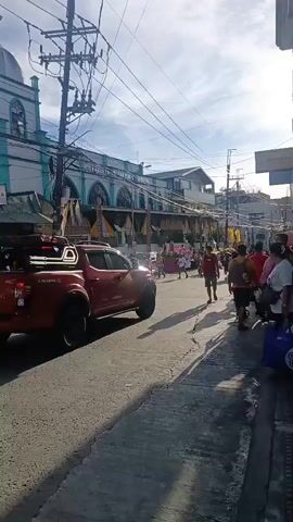 The video shows a street scene with a red pickup truck driving down the road. On the left side of the road is a church with a green and white facade, and what appears to be a procession or celebration taking place in front of it. There are people walking on the sidewalk on the right side of the road, some carrying bags and others pushing carts. The sky is bright with some clouds, and there are overhead power lines visible. The overall atmosphere appears to be festive.