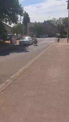 The video shows a street scene with a sidewalk and a road. A person on a bicycle is visible on the road, approaching a crosswalk. There are cars parked on the side of the road and buildings on either side of the street. The sky is blue with some clouds, indicating daytime. The scene appears to be in a residential or commercial area with trees and greenery.