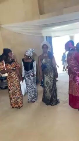 Four women dressed in traditional clothing are dancing together in a room with a simple decor. The room has light-colored walls, a ceiling draped with white fabric, and an open doorway leading to an area with more people. The women are all smiling and appear to be enjoying themselves. They are spaced apart from each other, but clearly dancing in coordination with the music. The clothing is distinctively patterned and vibrant, each woman wearing a unique design.