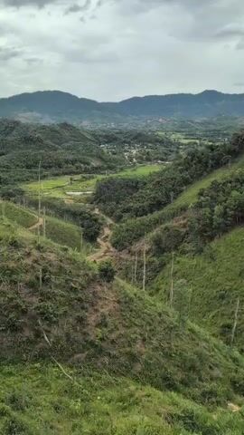 The video shows a panoramic view of a lush green valley with rolling hills and mountains in the background. The sky is cloudy and overcast. In the foreground, there are steep, grassy slopes with scattered trees and some areas of cleared land. A dirt path winds its way down one of the slopes. In the middle ground, there is a village or settlement nestled in the valley, surrounded by green fields and more trees. The distant mountains are covered in dense vegetation. The overall impression is one of natural beauty and a rural landscape.