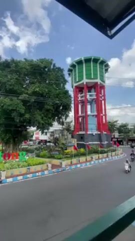 The video shows a water tower on a traffic circle with some passing motorcycles. A man is looking at his phone at a table and a young child appears to be playing with something.