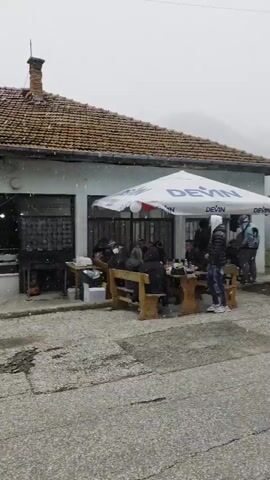 The video depicts a street scene on a cloudy day with light snow falling. A low building with a tiled roof and large window openings is visible on the right side of the street, with people seated at wooden tables under white umbrellas. Some people are walking in the street, and cars are parked further down the road. The street surface is cracked and uneven. Overhead power lines are also visible. A mournful male a cappella song is audible in the background.