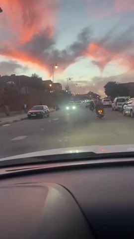 A vehicle is moving along a road. The sky is at dusk and is colored with orange and pink clouds. There are other vehicles on the road. A person is walking on the sidewalk along a roadside embankment. There are utility poles and streetlights. The vehicle continues to travel.
