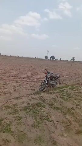 The video pans across an open, dry, and dusty field under a cloudy sky. A red motorcycle is parked in the field, leaning on its kickstand. In the distance, a few trees and what appears to be a power line tower are visible. The camera then shifts to show two people in the field. One person, wearing a red shirt, stands over another person who is sitting on the ground. They appear to be interacting, but the exact nature of their interaction is unclear. The scene is set in a rural, arid environment.