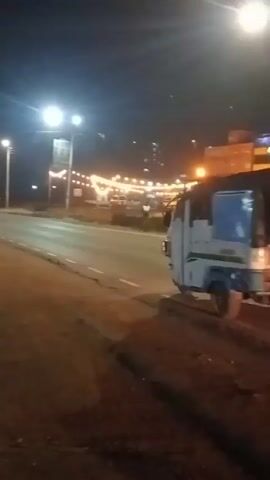The video shows a dark roadside scene at night. Streetlights illuminate the road and nearby buildings. Initially, a small truck with a dark load on its bed drives by. Later, a white minivan and a white sedan pass the camera. The camera appears to be stationary, capturing vehicles driving on the road and some adjacent dirt. The buildings in the background are adorned with festive lights.