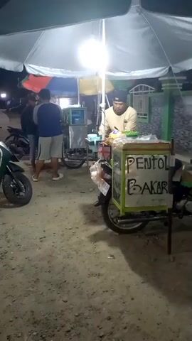 A street vendor sells grilled meatballs from a cart attached to a motorcycle under a large white umbrella. The vendor, wearing a light-colored long-sleeved shirt and a black cap, is preparing food. In the background, two customers stand near another cart. The ground is covered in dirt and gravel. Motorcycles are parked nearby. The scene is illuminated by a bright light under the umbrella. The cart has a sign that reads "PENTOL BAKAR," which translates to "Grilled Meatballs."