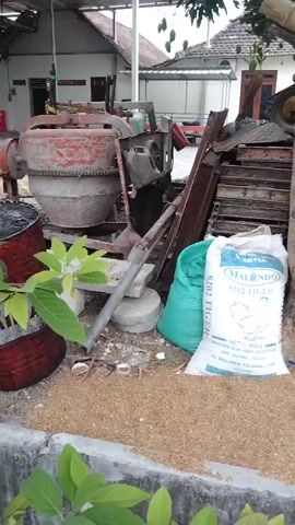 The video shows an outdoor scene with several objects piled up. In the foreground, there is a pile of light brown granular material. To the left of this material, there is a white bag with blue lettering that says "MALINDO 8202 TIGER". Next to the white bag is a large green bag. Behind these bags, there is a tree with a thick trunk and several branches, some of which are supported by bamboo poles. To the left of the tree, there is a red cement mixer and other metal objects. Further back, there are stacked orange crates, several tires, and what appears to be a building with a tiled roof. Green leaves from trees are visible in the upper part of the frame. The ground is uneven and appears to be dirt and gravel. The overall impression is one of a storage area or a construction site.