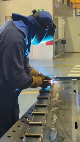 A person wearing a welding mask and protective clothing is welding a metal object on a workbench. Sparks are flying from the welding torch. The background shows a workshop or factory setting with other equipment and industrial elements.