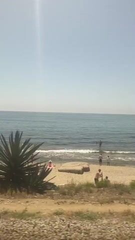 This video captures a sunny day at the beach in Mataró, Spain. The scene is a typical seaside resort with people enjoying leisure activities. The clear blue sky and calm sea suggest pleasant weather, likely midday given the brightness.

The foreground is a sandy beach littered with rocks and some sparse vegetation. Several beach umbrellas are set up, offering shade. People are scattered across the sand, some lounging under the umbrellas, others walking along the shoreline, and a few are in the shallow water. A couple of boats are visible in the distance on the water, indicating maritime activity. The overall atmosphere appears relaxed and leisurely, characteristic of a summer day at the beach. There is no visible text in the media file.