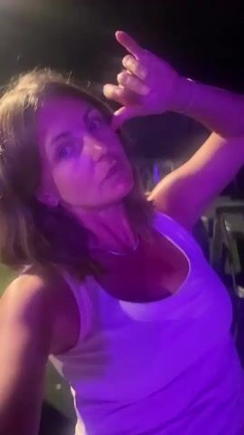 A woman is in front of the camera, lit by purple and pink lights, with her right arm extended upwards and her hand in a gesture. She appears to be speaking or singing, with her mouth open and her gaze directed at the camera. Her hair is brown and styled in loose waves, and she is wearing a white tank top and a thin silver necklace. The background is blurry and appears to be outdoors at night, with dark shapes that could be chairs or structures visible. The overall impression is of a person at an event or performance.