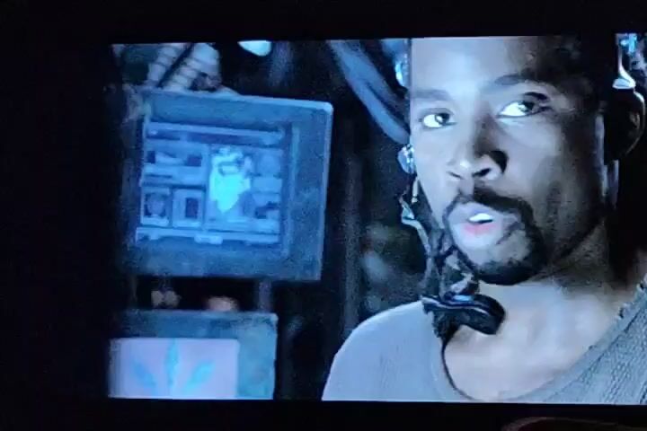 The video opens with a close-up of a Black man wearing headphones and a headset, looking towards the camera. He has a goatee and dark, braided hair. To his left, a monitor displays a blue interface with several windows containing text and images. The scene appears to be dimly lit, with the background out of focus.

The video then transitions to a shot of a man in sunglasses, with a bald head, wearing a black leather jacket. He is holding a dark object, possibly a bottle, to his mouth. He is inside a vehicle, as indicated by the car door and steering wheel visible in the frame.

Following this, the camera focuses on a woman also wearing sunglasses and a black outfit. She is also inside a vehicle and appears to be speaking. Her mouth is open, and her expression suggests she is actively communicating. The steering wheel is prominently visible in front of her. The lighting in this segment is brighter than the initial shot.