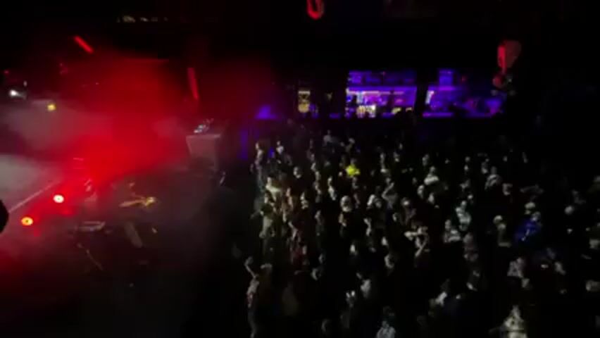 A live concert is underway. Red stage lights illuminate the performing band on the left side, partially obscured by a cloud of stage fog. A large crowd of people is visible, dancing and watching the performance. A bar area with lights is visible in the background.