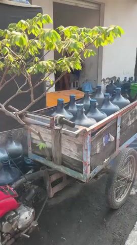 The video shows a man sitting in a doorway, possibly a shop or storage area. In front of the doorway, a cart is filled with large plastic water bottles with blue caps. More water bottles are visible stacked to the right of the doorway and a large blue cooler is partially visible on the left. The man is wearing a blue shirt and shorts. He appears to be looking at something on a table in front of him. The ground is rough and uneven, with some debris and puddles. The building appears to be made of concrete. The lighting suggests it is daytime, possibly outdoors or in a covered area.
