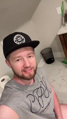A man wearing a gray t-shirt and a black baseball cap with a white logo is sitting on a toilet and recording a video of himself. He is smiling and looking at the camera. The bathroom has white marble floors and a white toilet. There are rolls of toilet paper on a shelf behind him and a black trash can in the corner.