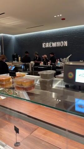 The video shows a food service establishment, likely a deli or prepared foods section of a grocery store. The camera pans across various food items displayed in glass cases. In the background, several employees are visible behind the counter, interacting with customers or preparing food. The environment appears clean and well-lit, with a modern aesthetic suggested by the dark wall behind the counter with white lettering that reads "BREW HOUSE."

Specifically, the video captures:
- In the foreground, glass display cases filled with various salads, prepared meals, and what appears to be seafood like salmon.
- Among the salads, there are dishes featuring chopped vegetables, grains, leafy greens, and what could be fruit salads.
- In one section of the display, there are containers of prepared food items.
- Behind the counter, several employees in casual attire are visible. Some are facing the camera, while others are turned away, possibly serving customers or working on food preparation.
- The back wall has a prominent logo or name of the establishment.
- In the middle ground, there are clear plastic containers with food items.
- The lighting is bright, illuminating the food displays and the work area.
- The overall impression is of a busy, organized food service environment.
