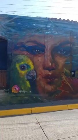 The camera pans across a street lined with buildings adorned with murals. The first building is painted blue and features a large mural of a face and a parrot.  Next to it, the building is red with a mural of a winged heart. Further down the street, buildings and walls are painted blue and also feature murals.  A yellow curb runs along the edge of the street. In the background, a mountain is visible.